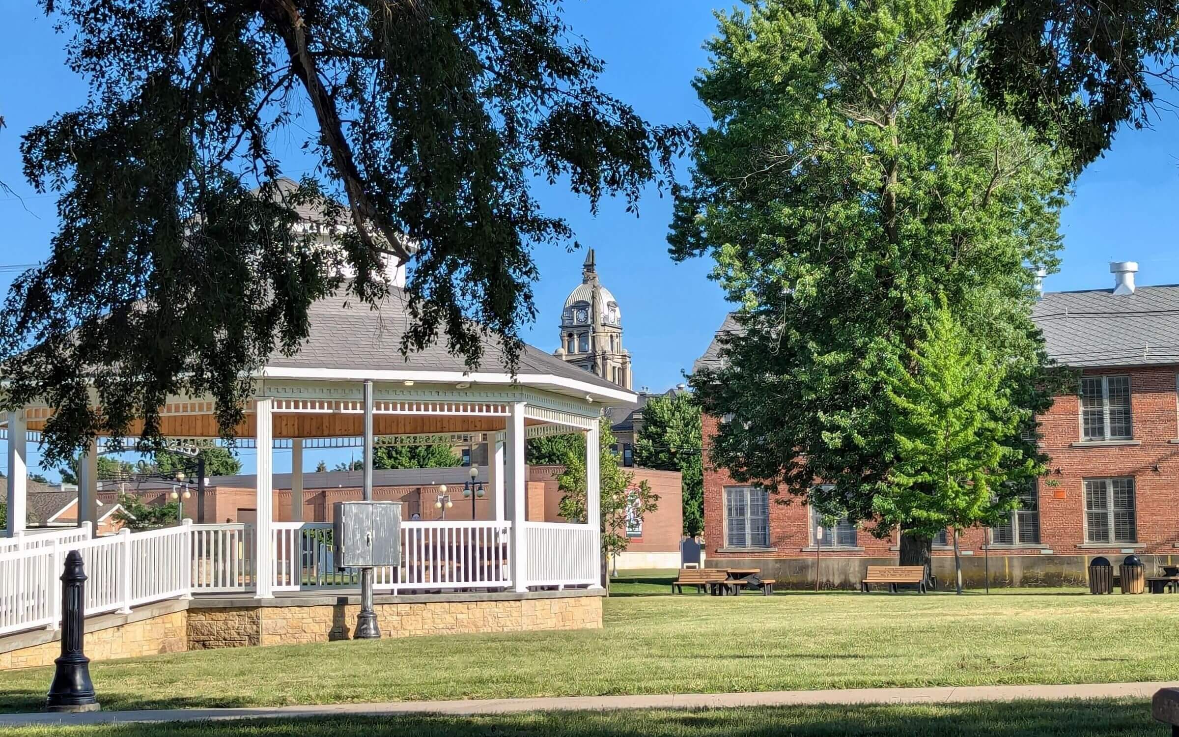 A white gazebo in a grassy park area with a lamppost, trees, and brick buildings in the background under a clear blue sky.