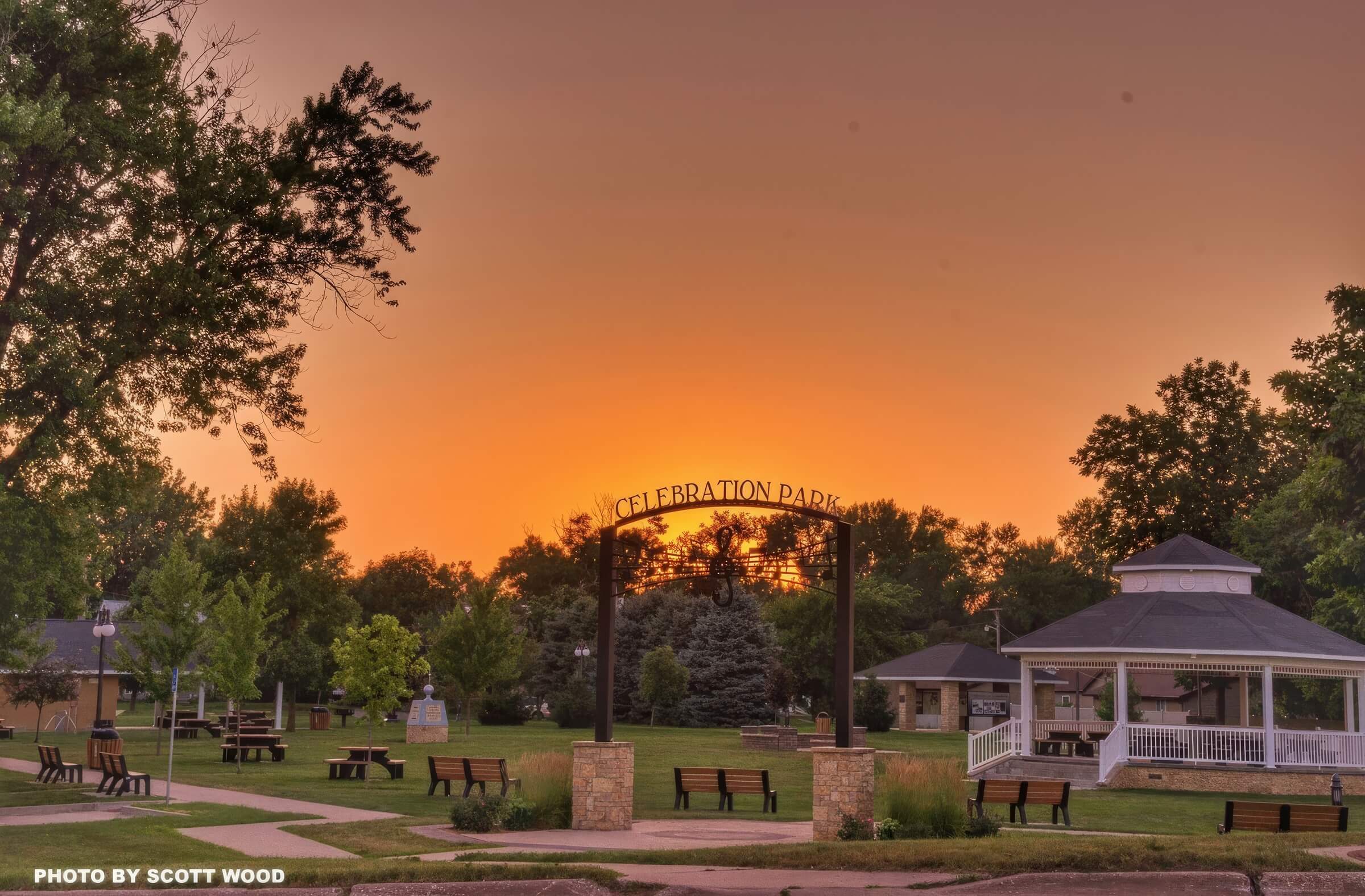 Celebration Park at sunset with a gazebo on the right, an archway sign in the center, and several benches in a grassy area. Trees and a house are visible in the background.