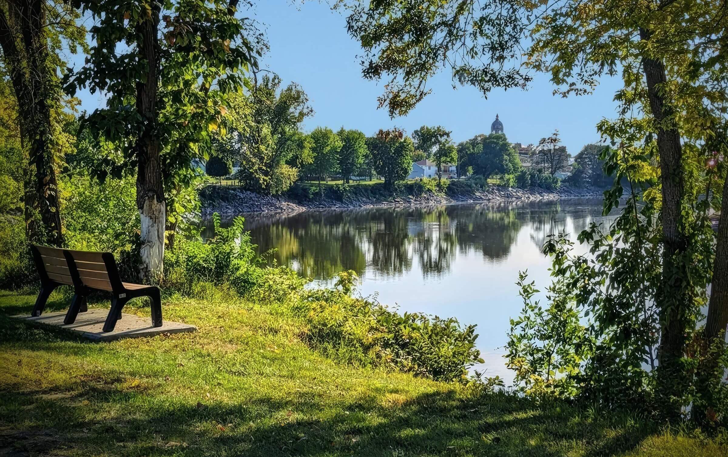 A wooden bench on a grassy area beside a calm river, surrounded by trees with a view of a park and distant building under a clear blue sky.