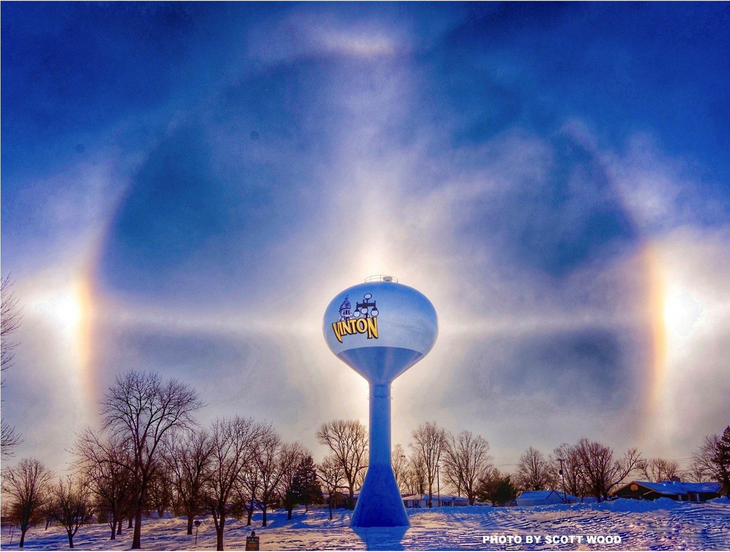 Water tower labeled "Vinton" with a bright halo and sundogs in the sky, surrounded by bare trees and snow on the ground.